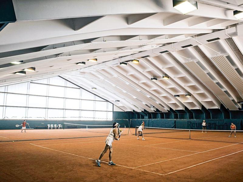 Indoor-Tennisplatz mit rotem Sandboden, großen Fenstern und spielenden Personen in Sportkleidung.