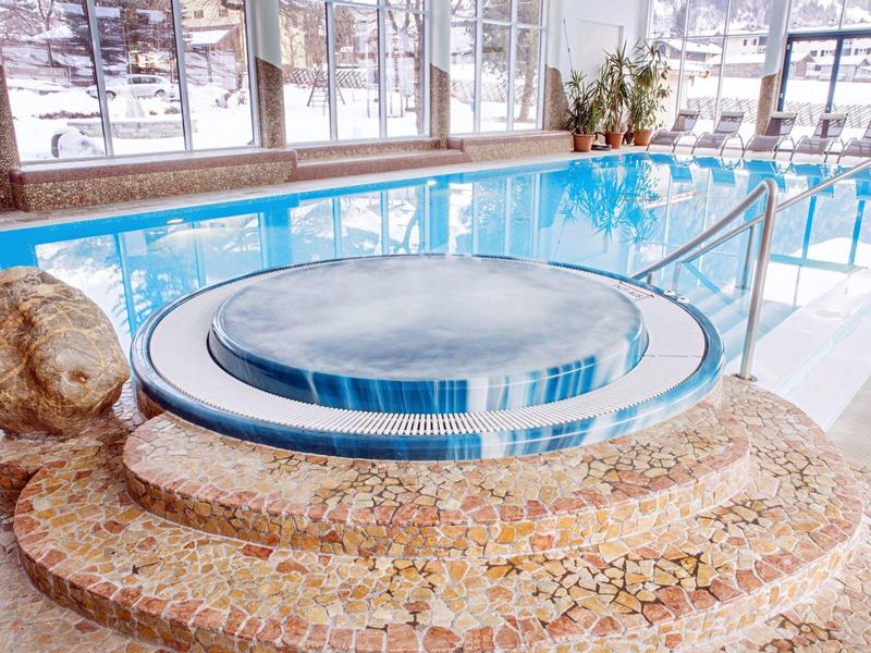 Runder Whirlpool mit blauer Umrandung in hellem Raum neben großem Schwimmbecken mit Sitzstufen.