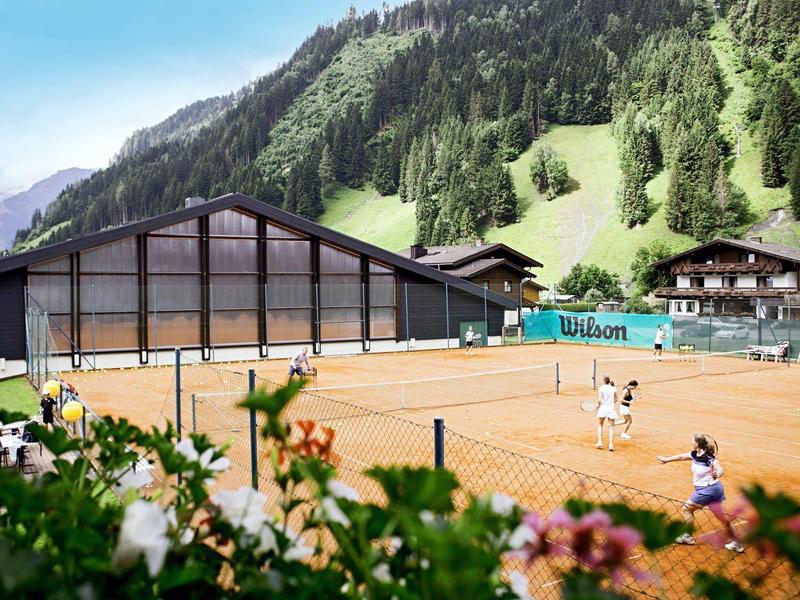 Zwei Personen spielen Tennis auf einem Sandplatz in einer Berglandschaft mit einer großen Holzhütte.