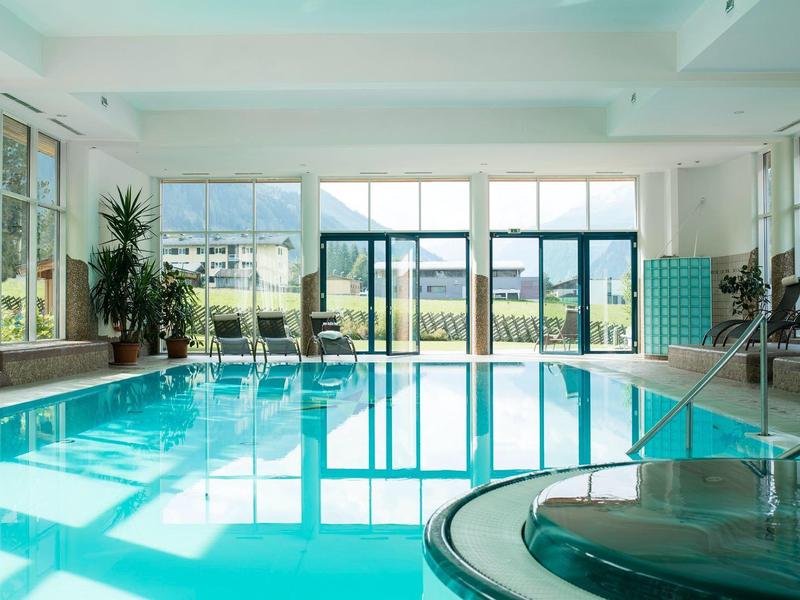 Heller Innenpool mit Glaswand, Pflanzen links und Liegestühlen rechts, Blick nach draußen.