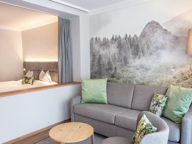 Gemütliche Sitzecke mit grauem Sofa, grünen Kissen und Wald-Wandbild in hellem Raum.