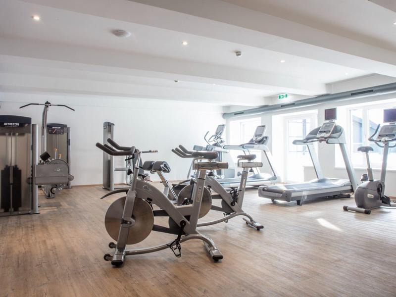 Moderner Fitnessraum mit Fahrradergometer, Laufbändern und Kraftgeräten, heller Raum mit Holzboden.