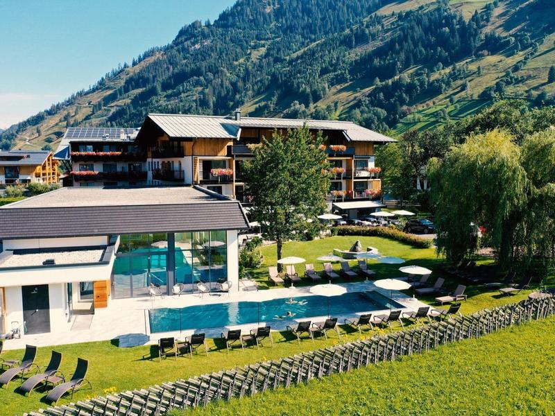 Bergdorf mit modernen Chalets, Außenpool und Liegestühlen vor bewaldeten Alpenhängen.