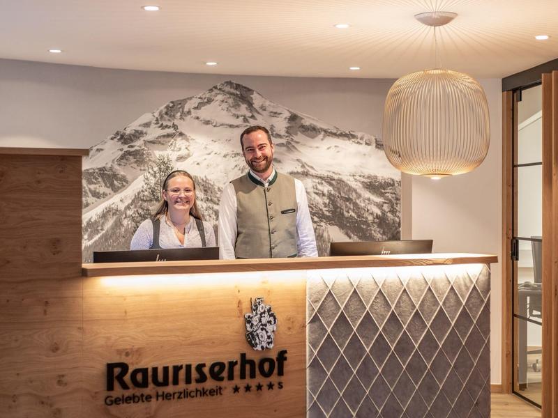 Rezeption mit Holztheke, zwei freundlich lächelnden Mitarbeitern und Bergfoto an der Wand.