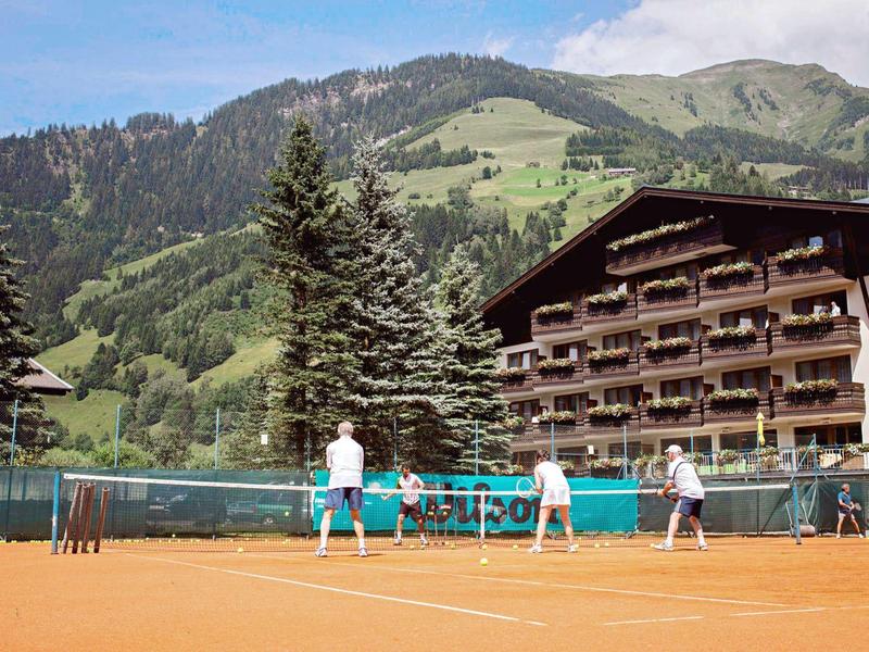 Tennisspieler auf rotem Sandplatz vor bewaldetem Berg und mehrstöckigem Holzgebäude.
