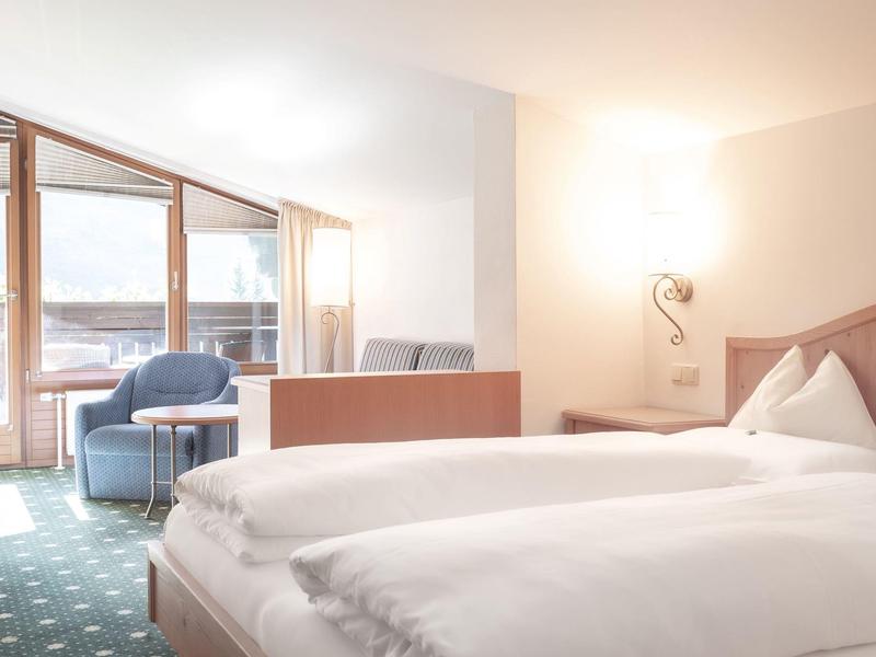 Helles Hotelzimmer mit Doppelbett, Sesseln und großem Fenster mit Blick auf verschneite Berge.