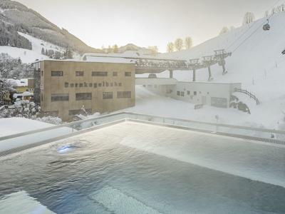 Podgrzewany odkryty basen w zaśnieżonym górskim krajobrazie obok budynków hotelowych o porannym świetle.