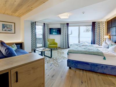 Jasny pokój hotelowy z dużym łóżkiem, miejscem do siedzenia, telewizorem i balkonem z widokiem na góry.