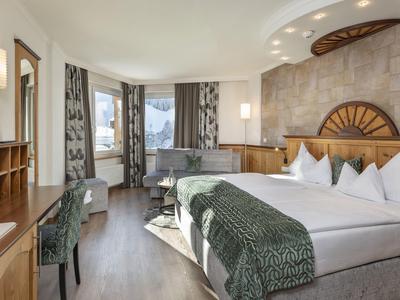 Jasny pokój hotelowy z dużym łóżkiem, biurkiem, krzesłem i panoramicznymi oknami z widokiem na góry.