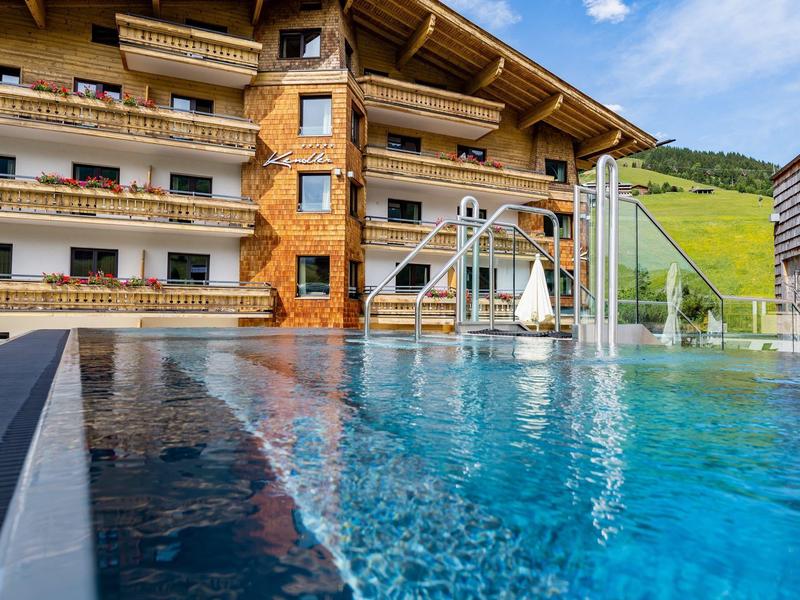 Hotel alpejski z basenem na świeżym powietrzu, drewnianą fasadą i zielonymi wzgórzami w tle pod niebieskim niebem