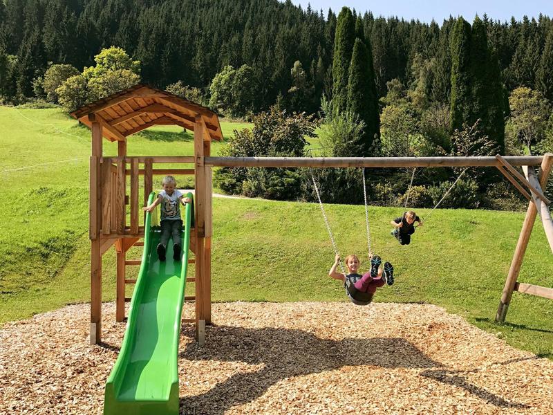 Aire de jeux pour enfants avec toboggan vert et balançoire devant une colline boisée.