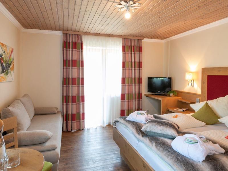 Chambre d'hôtel confortable avec lit double, canapé, télévision, balcon, plafond en bois et couleurs chaudes.