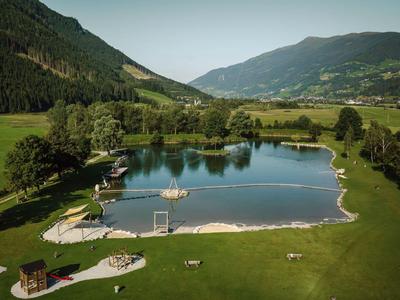 Klarer Badesee inmitten grüner Wiesen mit umliegenden Bergen und bewaldeten Hängen.