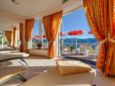 Jasny pokój relaksacyjny z leżakami i pomarańczowymi zasłonami, widok na taras z czerwonymi parasolami i jezioro.