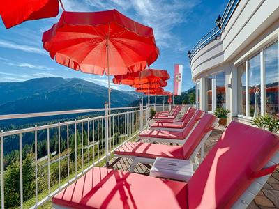 Rode ligstoelen en parasols op een hotelterras met uitzicht op bergen.