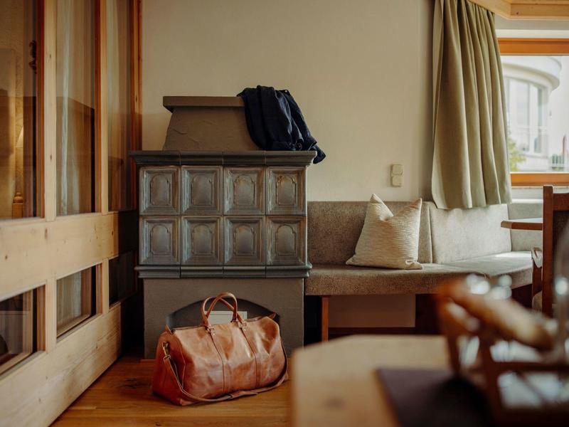 Przytulny kąt hotelowy z kaflowym piecem, drewnianymi meblami i torbą podróżną na podłodze.