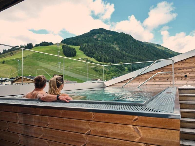 Two people relax in an outdoor pool with mountain views and wooden enclosure.