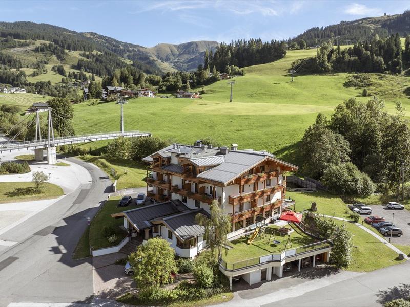 A large mountain hotel with balcony terraces in front of green hills and forested mountains.