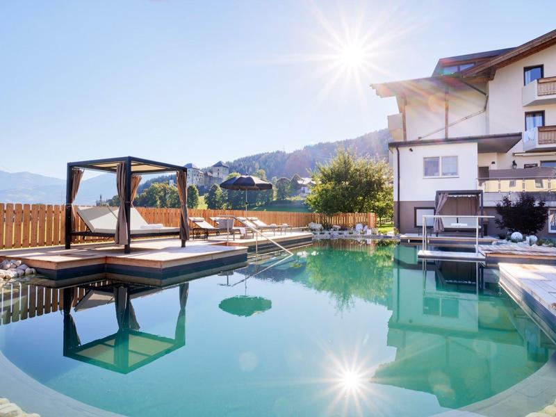 Moderner Außenpool mit Holzdeck und Lounge-Bereich vor ländlichem Haus, Berge im Hintergrund, sonnig.