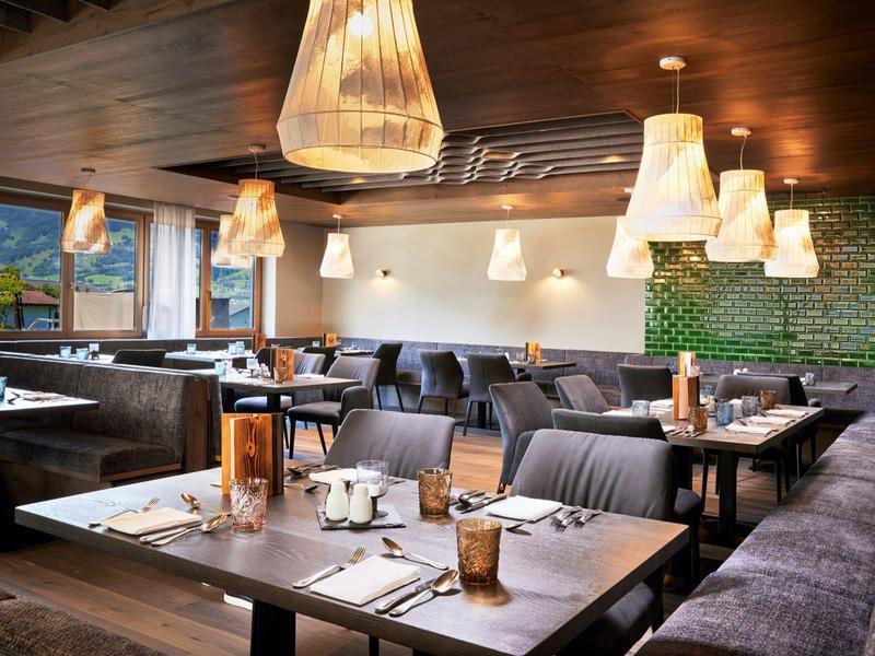 Modernes Restaurant mit Holztischen, braunen Stühlen, grün gefliester Wand und warmen Hängelampen.