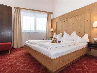 Jasny pokój hotelowy z drewnianym łóżkiem, czerwonym dywanem i dużym oknem z zasłonami.