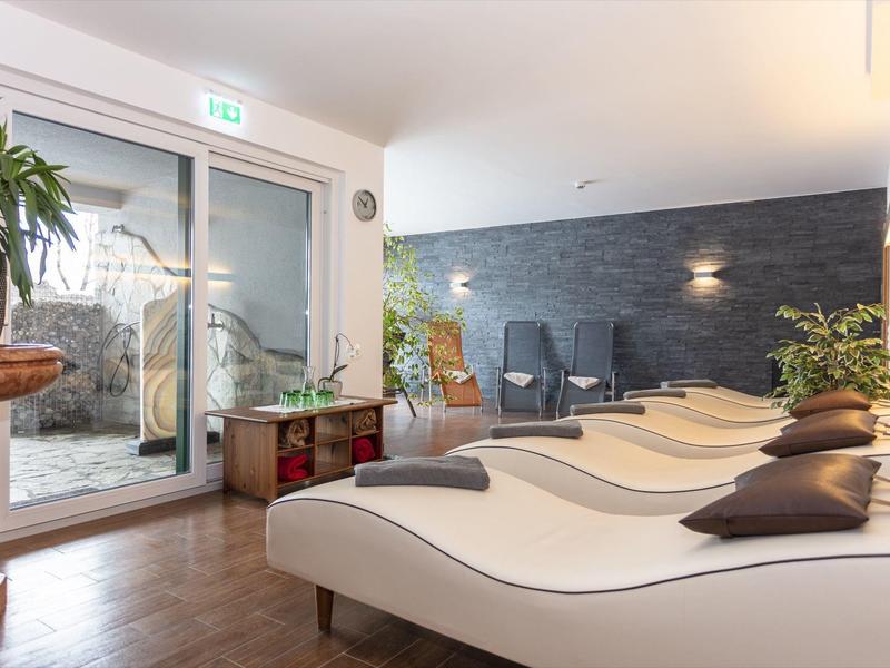 Pokój relaksacyjny z leżakami, poduszkami, dwoma fotelami i roślinami przy szklanych drzwiach.