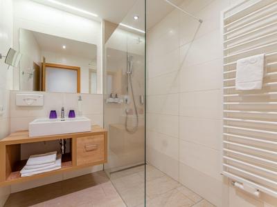 Moderní koupelna se skleněným sprchovým koutem, dřevěnou umyvadlovou skříňkou a bílými ručníky.