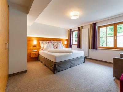 Světý hotelový pokoj s velkou postelí, kobercem a velkým oknem s výhledem na zeleň.