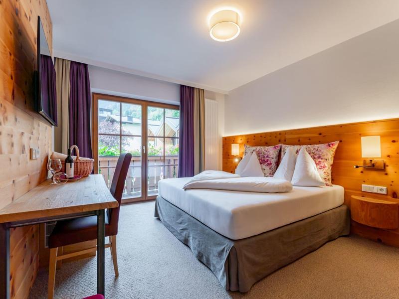 Moderní hotelový pokoj s manželskou postelí, dřevěnými prvky a výhledem z balkonu na zelenou krajinu.