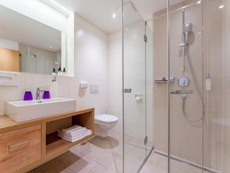 Jasná koupelna se skleněnou sprchou, dřevěnou umyvadlovou skříňkou a bílým záchodem.