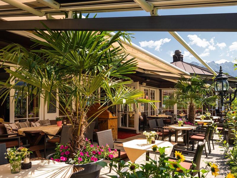 Słoneczny taras hotelowy z stołami, krzesłami i tropikalnymi roślinami.