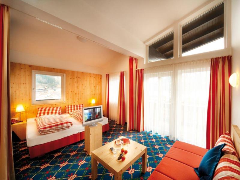 Przytulny pokój hotelowy z łóżkiem podwójnym, telewizorem i częścią wypoczynkową, jasne zasłony i kolorowe dywany.