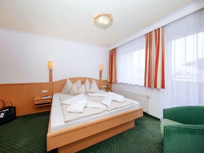 Světle zařízený hotelový pokoj s manželskou postelí, zeleným kobercem a pruhovanými závěsy.