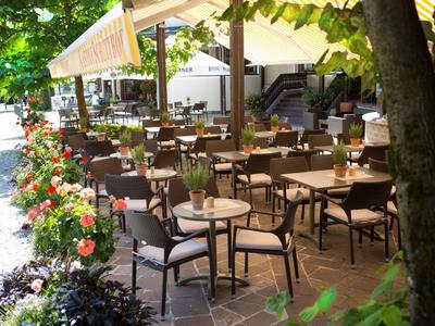 Terrazza all'aperto di un bar con molti tavoli e sedie sotto ombrelloni circondati da piante.