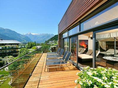 Balkon mit Holzterrasse, Liegestühlen und Glasgeländer vor Bergpanorama und blühenden Pflanzen.