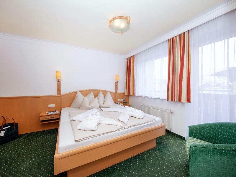 Světle zařízený hotelový pokoj s manželskou postelí, zeleným kobercem a pruhovanými závěsy.