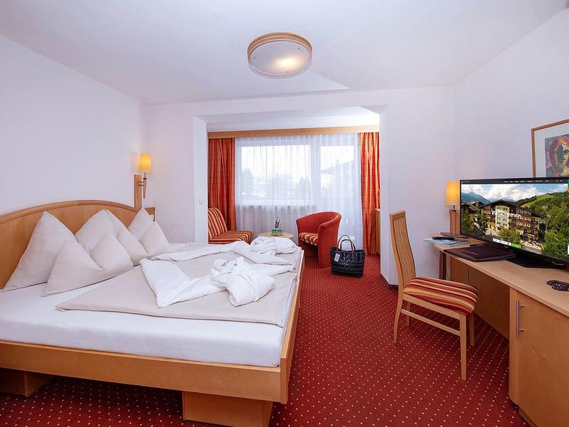 Světý hotelový pokoj s manželskou postelí, stolem, židlemi, televizí a oknem se závěsy.