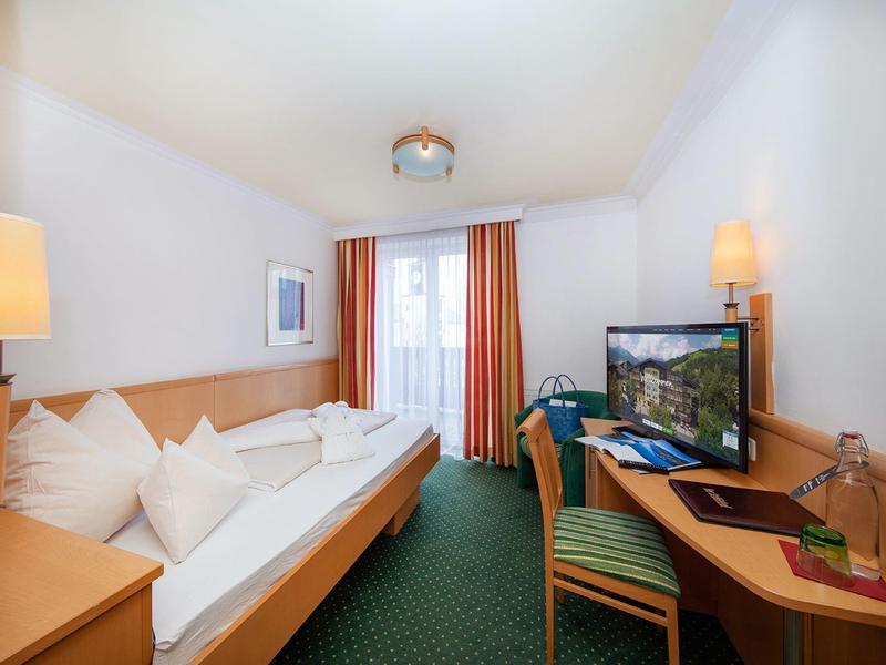 Světý hotelový pokoj se dvěma postelemi, psacím stolem, židlí a oknem se záclonami.