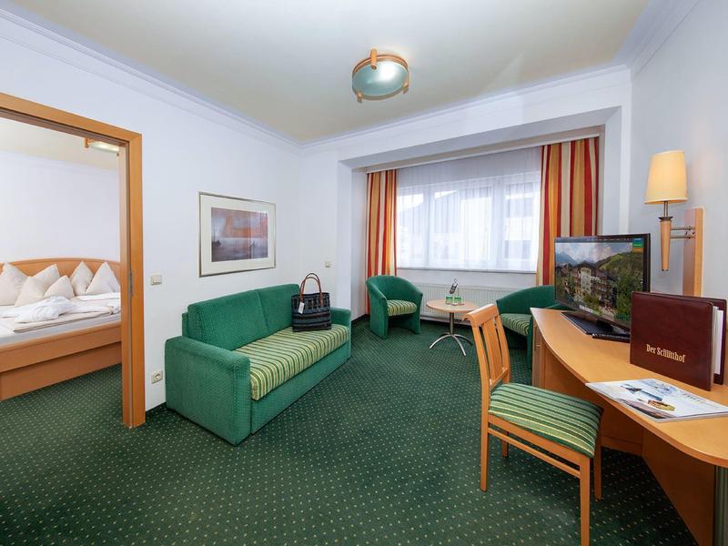 Světle hotelový pokoj se zeleným kobercem, pohovkou, stolem a přilehlou ložnicí.