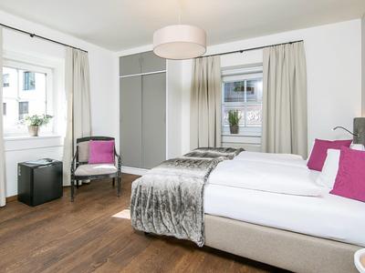 Modernes Schlafzimmer mit Holzboden, grauem Bett, pinken Kissen und großen Fenstern mit Gardinen.