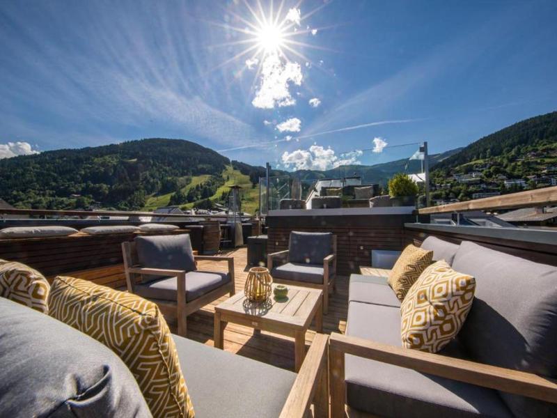 Gemütliche Terrasse mit Holzmöbeln, grauen Polstern, Kissen und Blick auf grüne Berge bei Sonnenschein.