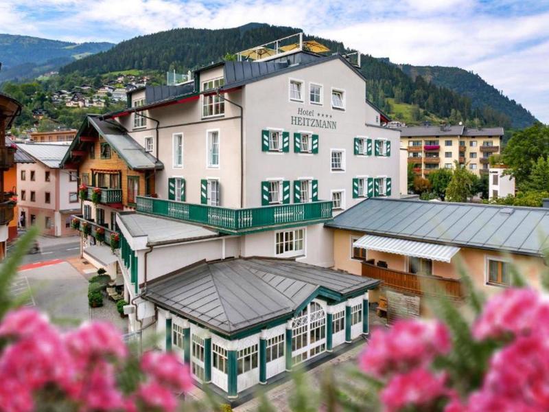 Buntes Dorf mit traditionellen Gebäuden vor grünen Bergen und blauem Himmel, rosa Blumen im Vordergrund.
