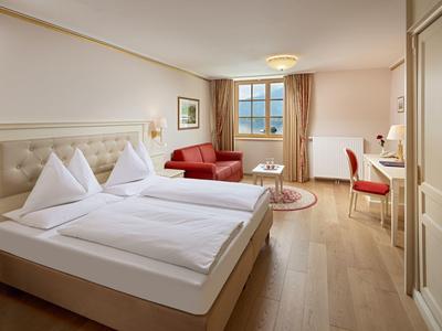 Světý hotelový pokoj s manželskou postelí, červenou pohovkou, psacím stolem a dřevěnou podlahou.