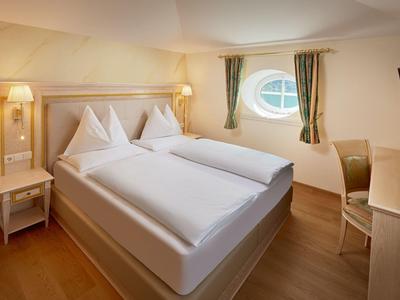 Světý hotelový pokoj se dvěma jednolůžky, dřevěnou podlahou a oválným oknem se zelenými závěsy.