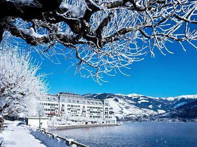 Zimní krajina se zasněženými stromy, jezerem a hotelem u hor pod jasně modrou oblohou