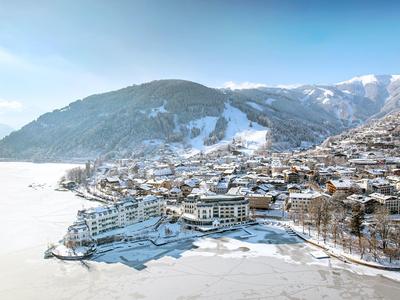 Zasněžená horská vesnice s zamrzlým jezerem a horami v pozadí pod jasnou oblohou