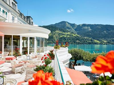 Hotelová terasa s červenými židlemi s výhledem na jezero a hory za slunečného dne.