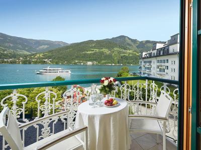 Balkon s stolem, židlemi a květinami s výhledem na jezero a zelené hory za čisté oblohy.