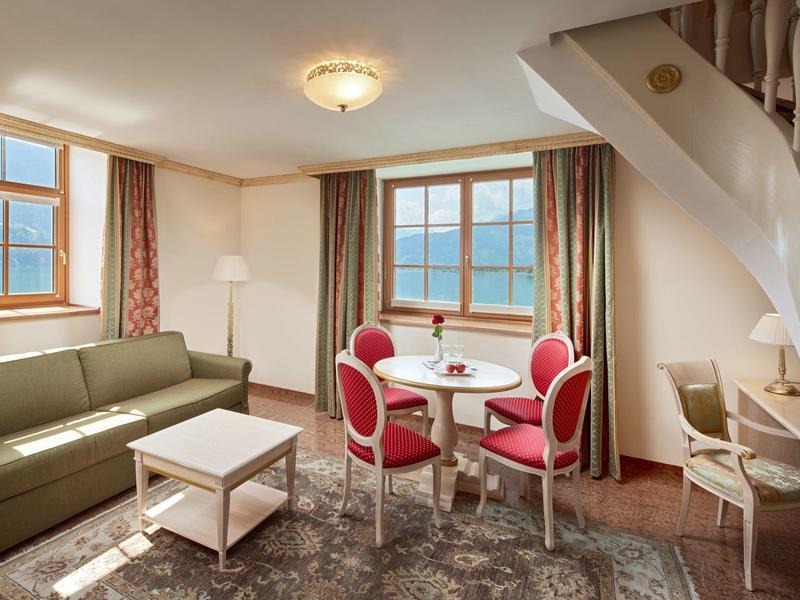 Příjemný hotelový obývací pokoj s pohovkou, malým stolkem a kulatým stolem se čtyřmi červenými židlemi.