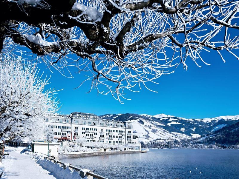 Zimní krajina se zasněženými stromy, jezerem a hotelem u hor pod jasně modrou oblohou